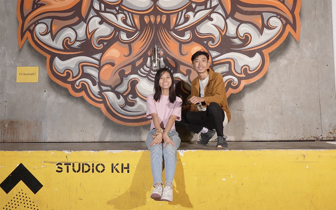 Two people sitting on a ledge in factory phnom penh with some kind of graffiti in the background and a 'Studio KH' sign on the wall./Image supplied by Joblink for Press Release.